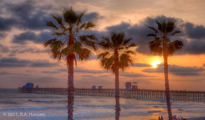 The warm, cloud-shrouded glow of the setting sun silhouettes the foreground palm trees and reflects off the water along the pier...