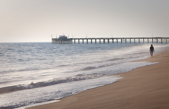 A lone walker strolls along the beach and surf, with the iconic pier in the background